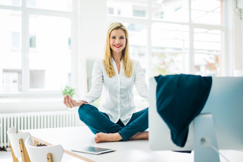 Smiling businesswoman sitting on desk in office practicing yoga